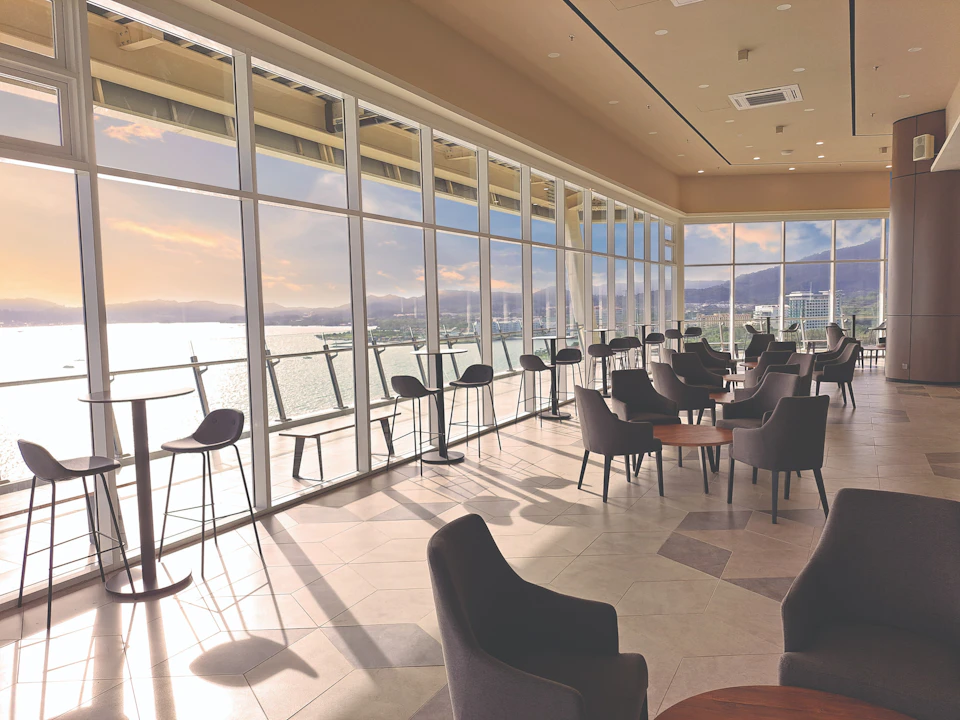 A modern cafe with large windows overlooking a waterfront view, featuring tables, chairs, and a bright, spacious interior.
