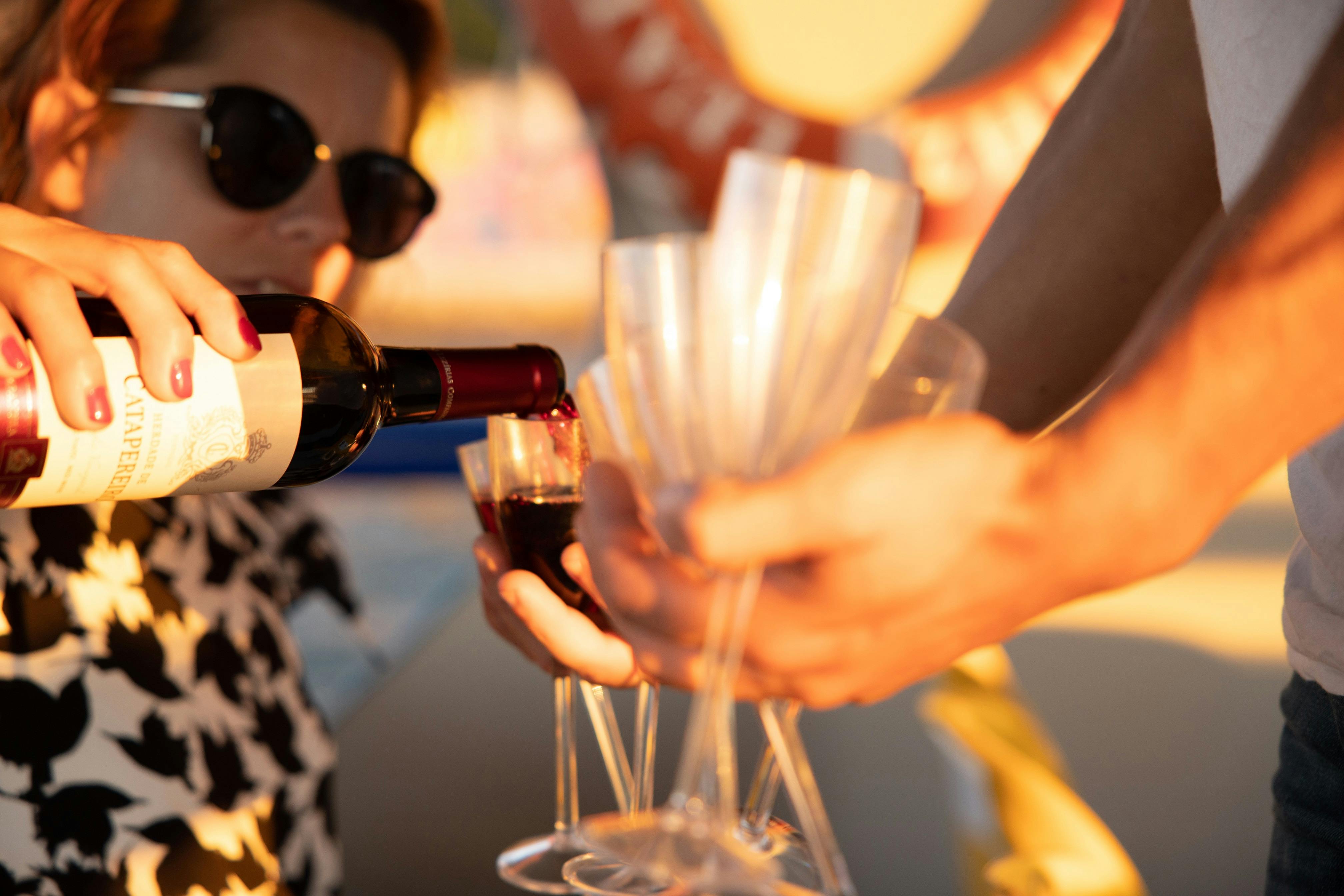 Persona con occhiali da sole che versa vino rosso da una bottiglia in più bicchieri trasparenti tenuti da qualcun altro.