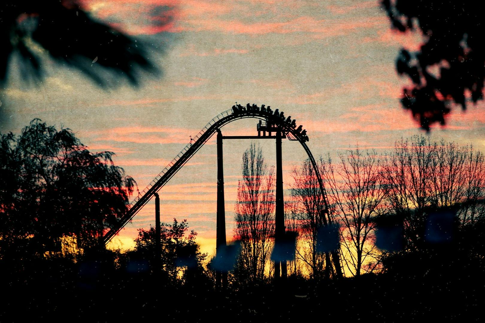 Horská dráha se siluetami jezdců proti zářivému západu slunce, obklopená stromy.