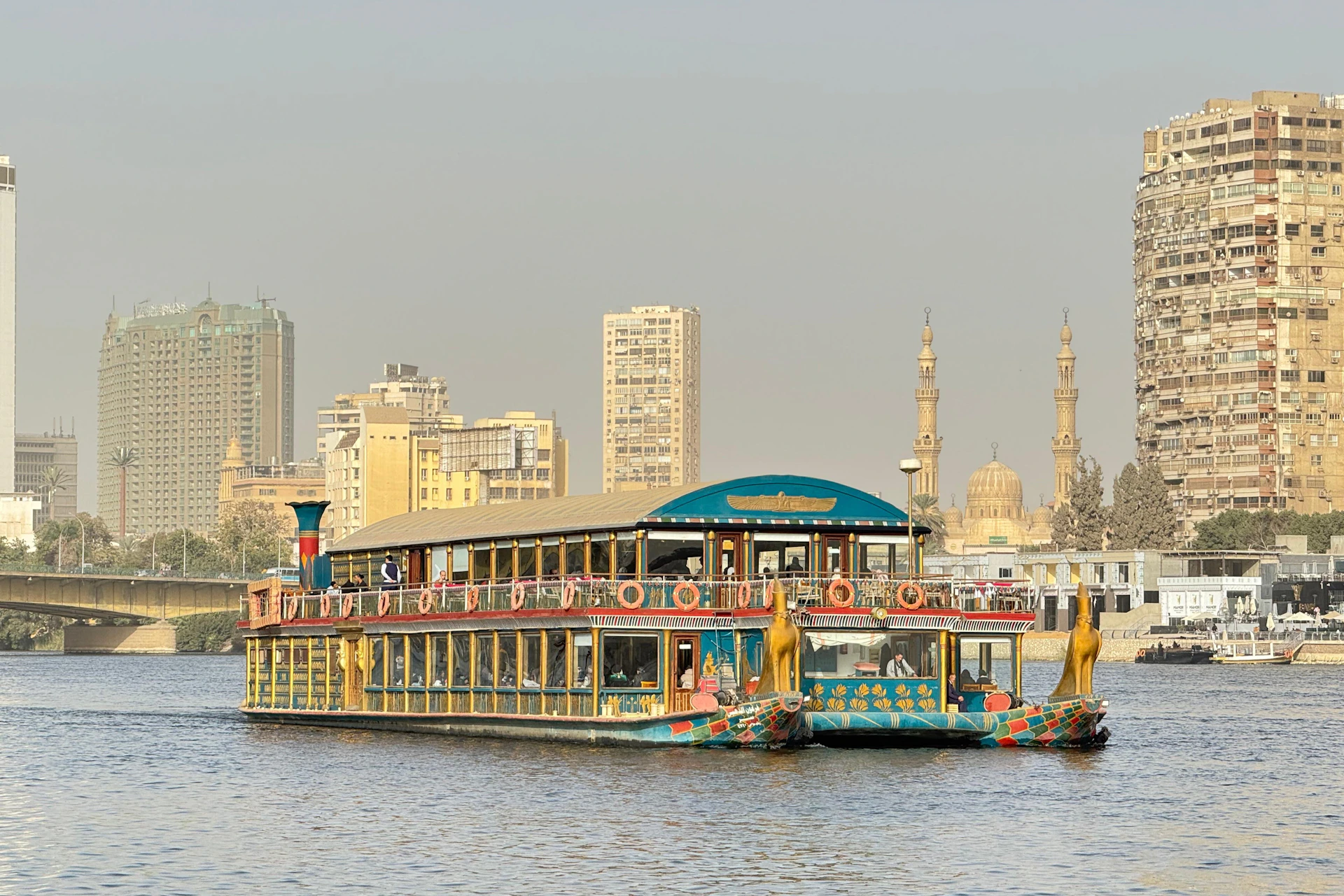 A riverboat styled with Egyptian motifs on a river, with a city skyline and two minarets in the background.