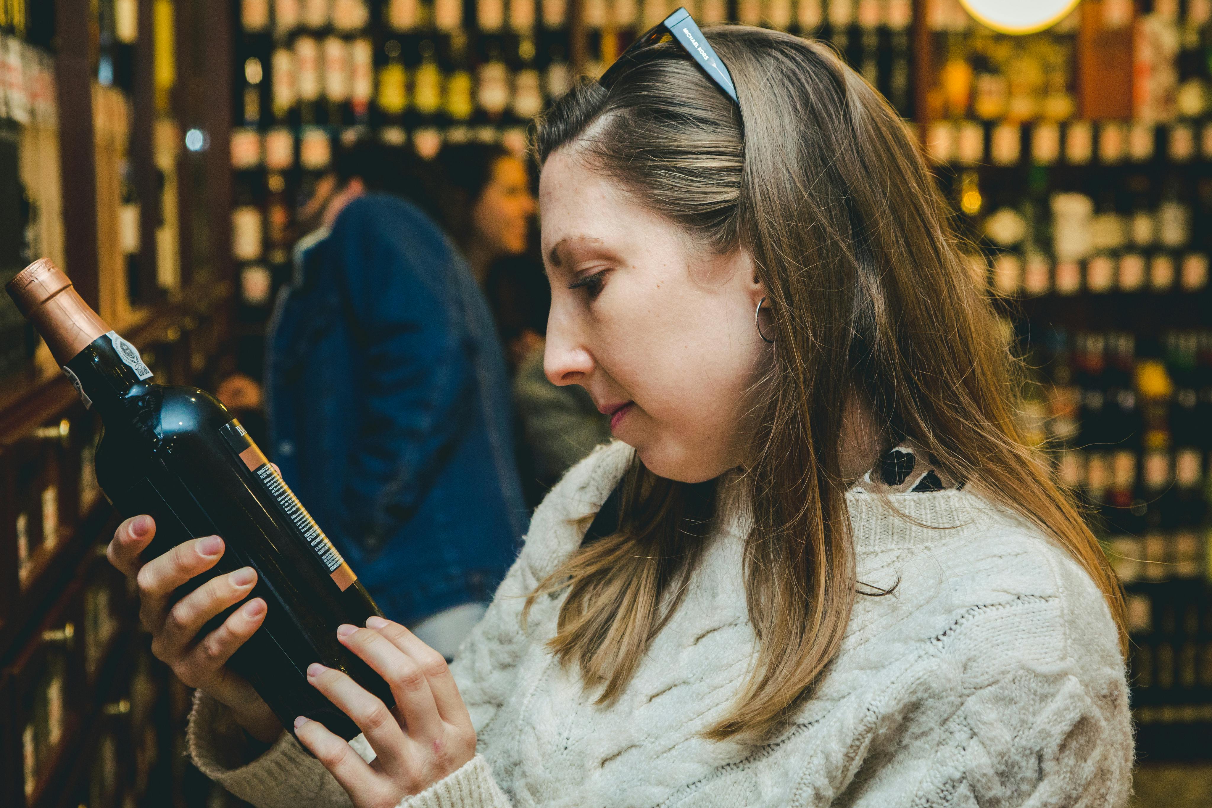 Een vrouw in een witte trui bekijkt een fles in een winkel met op de achtergrond rekken vol flessen.