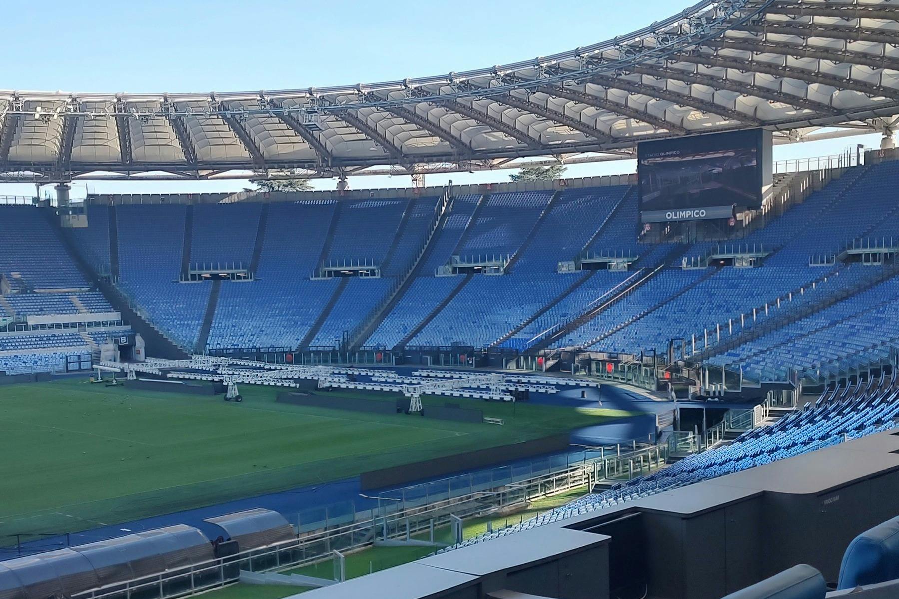 Een leeg stadion met blauwe stoelen, een groen veld en een grote dakconstructie boven je hoofd bij daglicht.