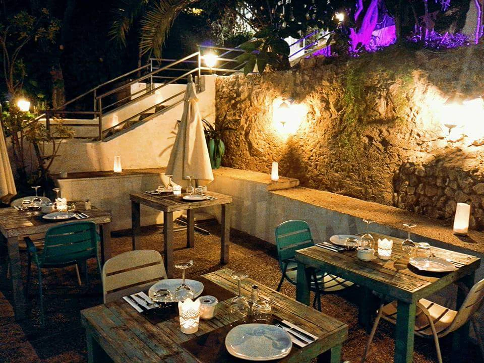 Outdoor restaurant seating area with wooden tables set for dinner, stairs, greenery, and warm lights creating a cozy ambiance.