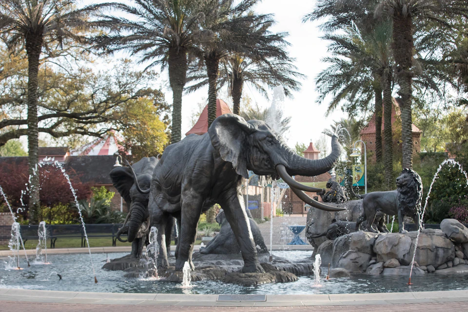 A fountain featuring sculptures of elephants surrounded by palm trees and buildings in an outdoor setting.