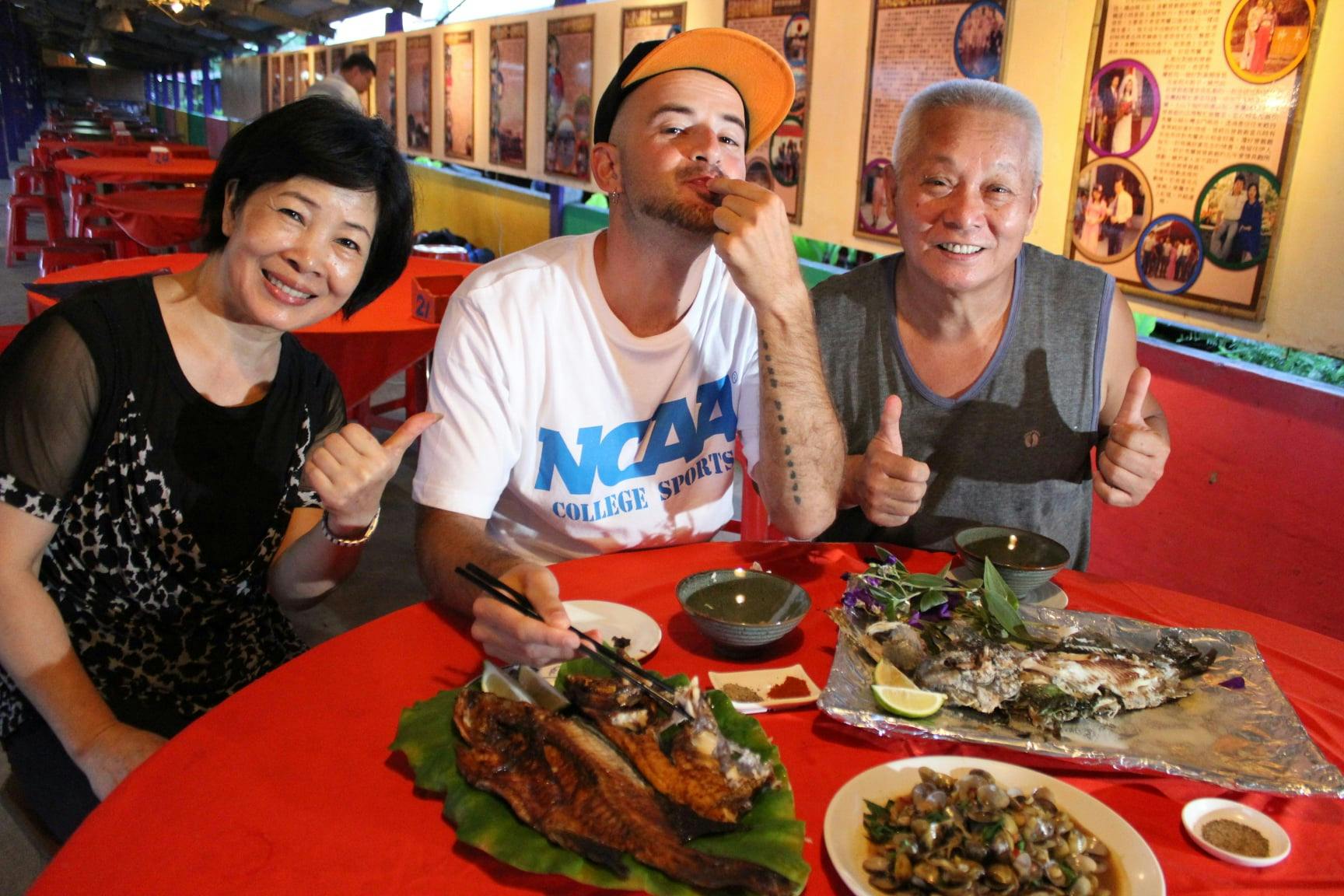 Three people sitting at a table with plates of food, smiling and gesturing, in a lively restaurant setting.