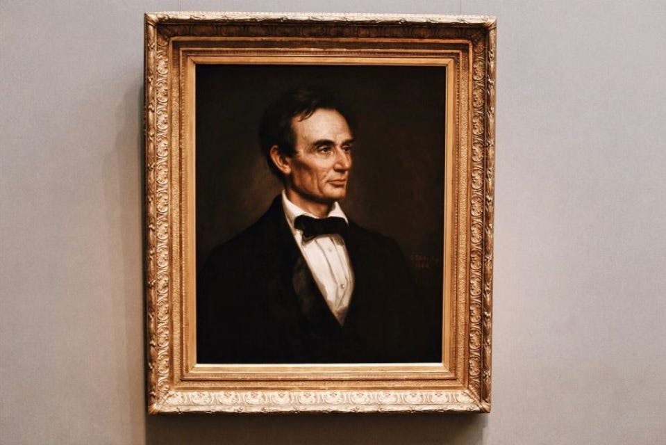 Ένα κορνιζαρισμένο πορτρέτο ενός άνδρα με επίσημη ενδυμασία, με σκούρα μαλλιά και παπιγιόν, που εμφανίζεται σε έναν γκρίζο τοίχο.