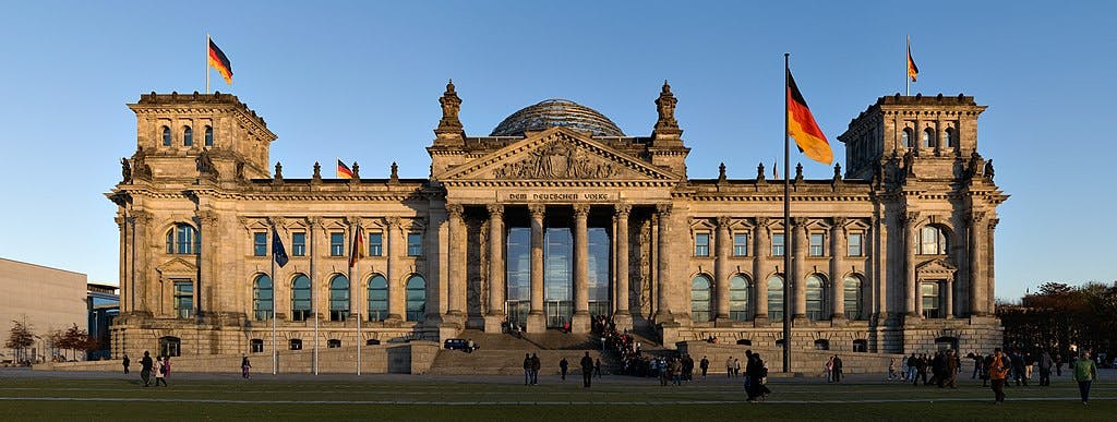 Large historic building with columns and a dome at the center, flying German flags, surrounded by people and green lawn.