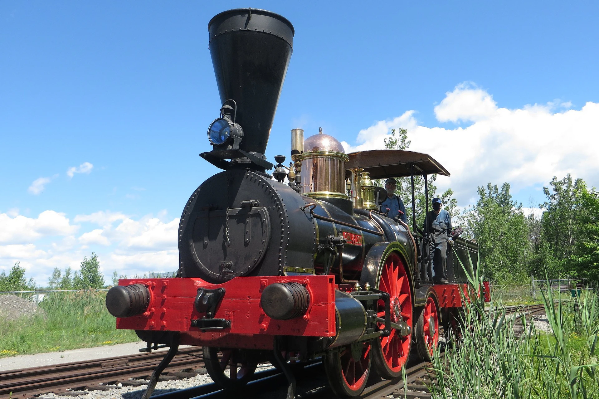 A vintage steam locomotive with red wheels and brass details travels on railroad tracks; two people are visible in the cab.