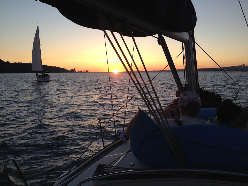 People on a sailboat at sunset with another sailboat visible in the distance on a calm body of water.