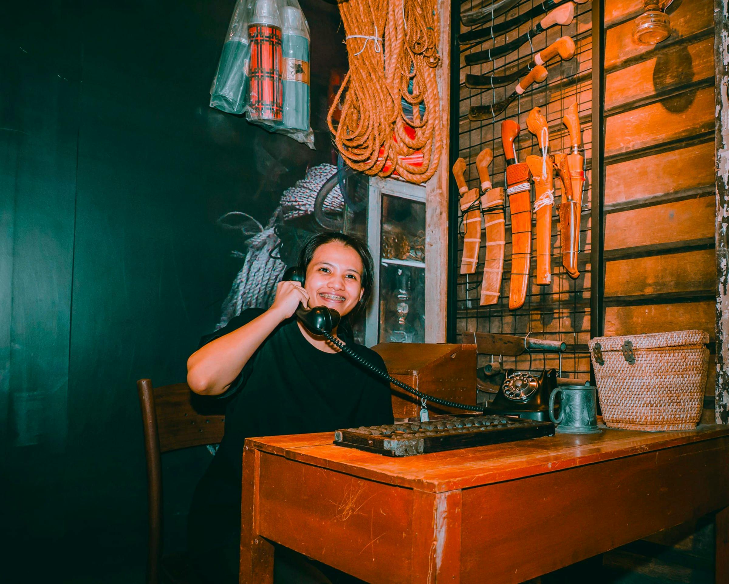 A woman smiling while holding an old-fashioned telephone in a rustic room with tools and ropes displayed on the wall.