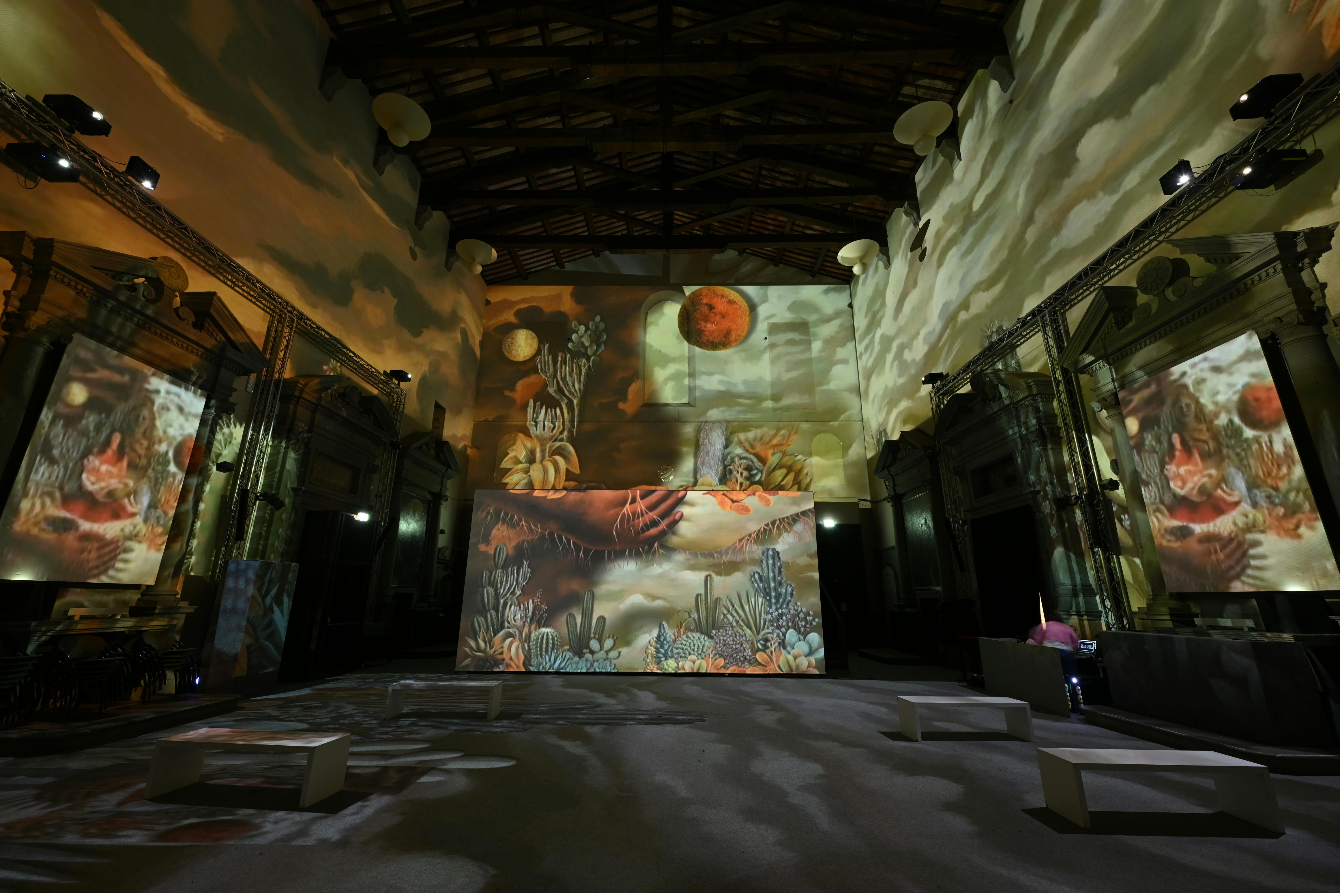 A large indoor space with desert-themed artwork and projections on walls, featuring cacti, a red moon, and benches.
