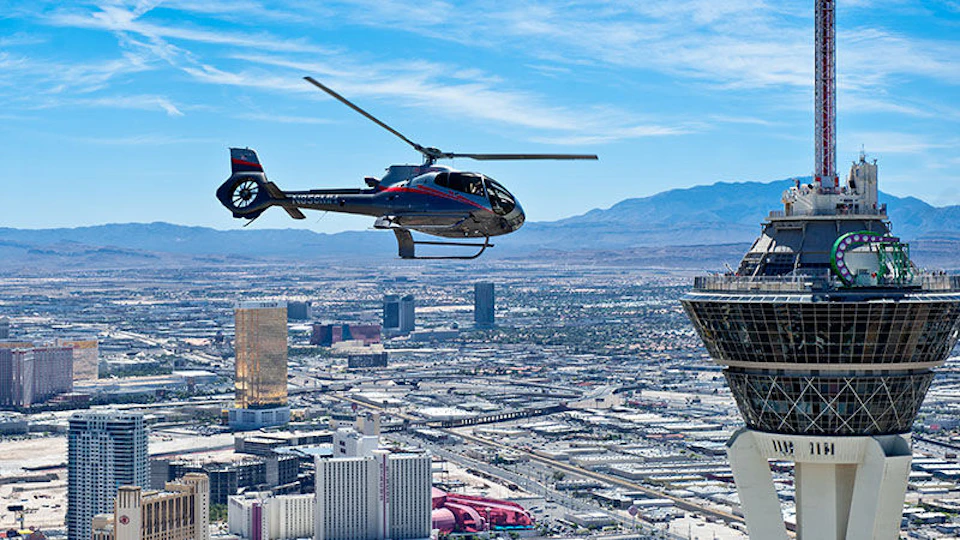 A helicopter flies over a cityscape with tall buildings, mountains in the background, and a large tower on the right.