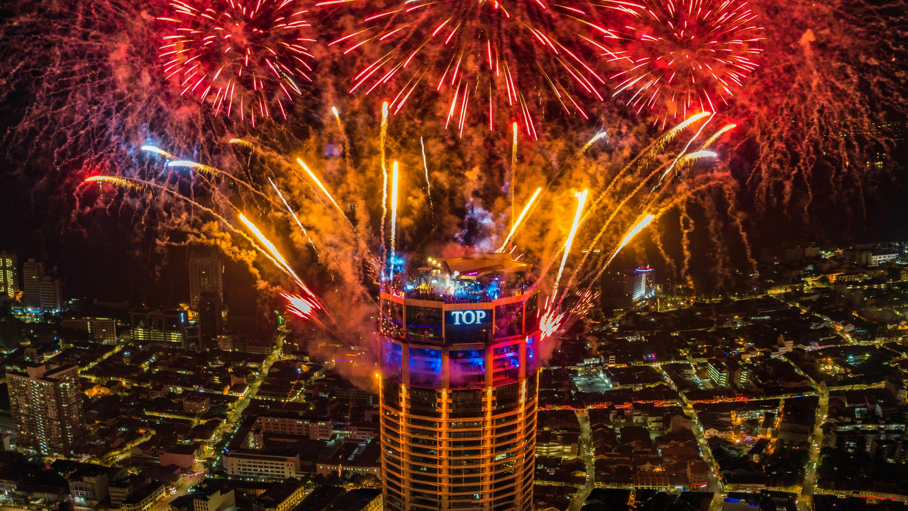Un grand immeuble, sur lequel est accrochée une enseigne lumineuse indiquant « TOP », est entouré de feux d'artifice aux couleurs éclatantes, avec en toile de fond le paysage urbain nocturne.