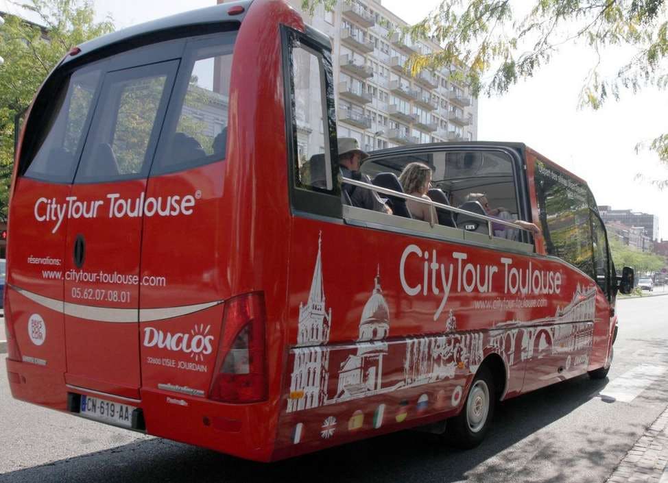 Autobus turistico rosso brillante con la scritta &#34;CityTour Toulouse&#34; con persone all&#39;interno, parcheggiato su una strada con edifici sullo sfondo.