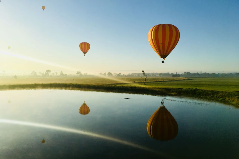 Two hot air balloons in the sky, reflected in a calm lake. One balloon is closer and larger, and the other is smaller in the distance.