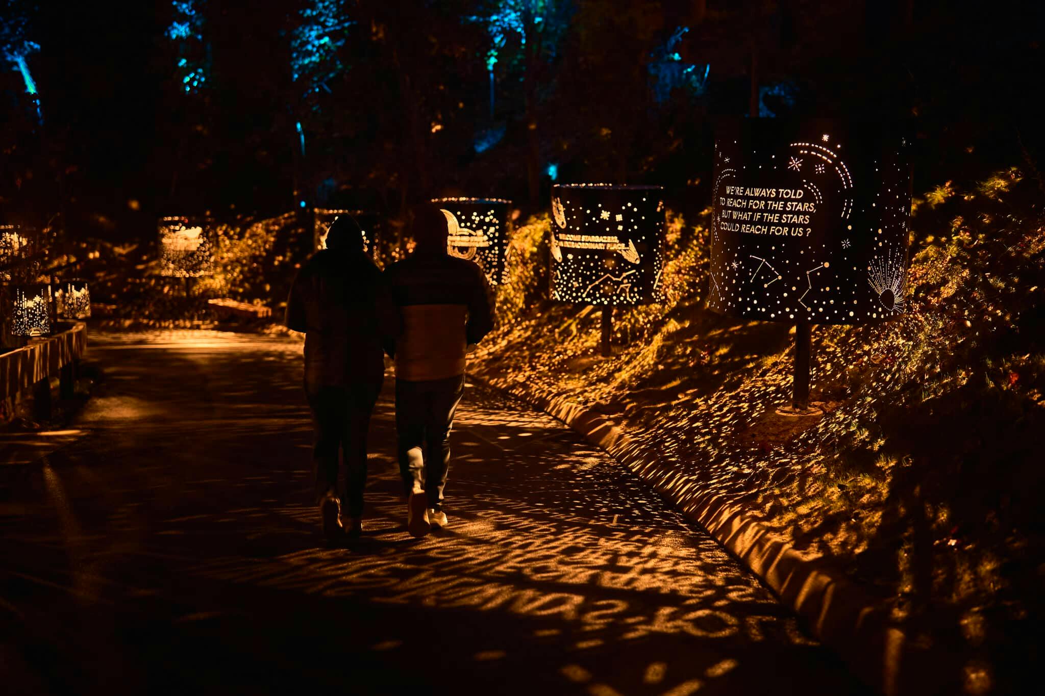 Пара идет по тропинке, освещенной художественными световыми узорами, проецируемыми из близлежащих декоративных панелей ночью.