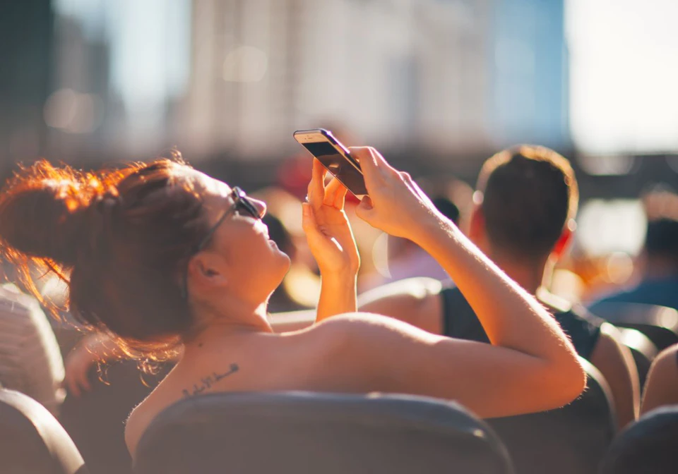 A person with sunglasses and a tattoo takes a photo with a smartphone in an outdoor setting with others in the background.