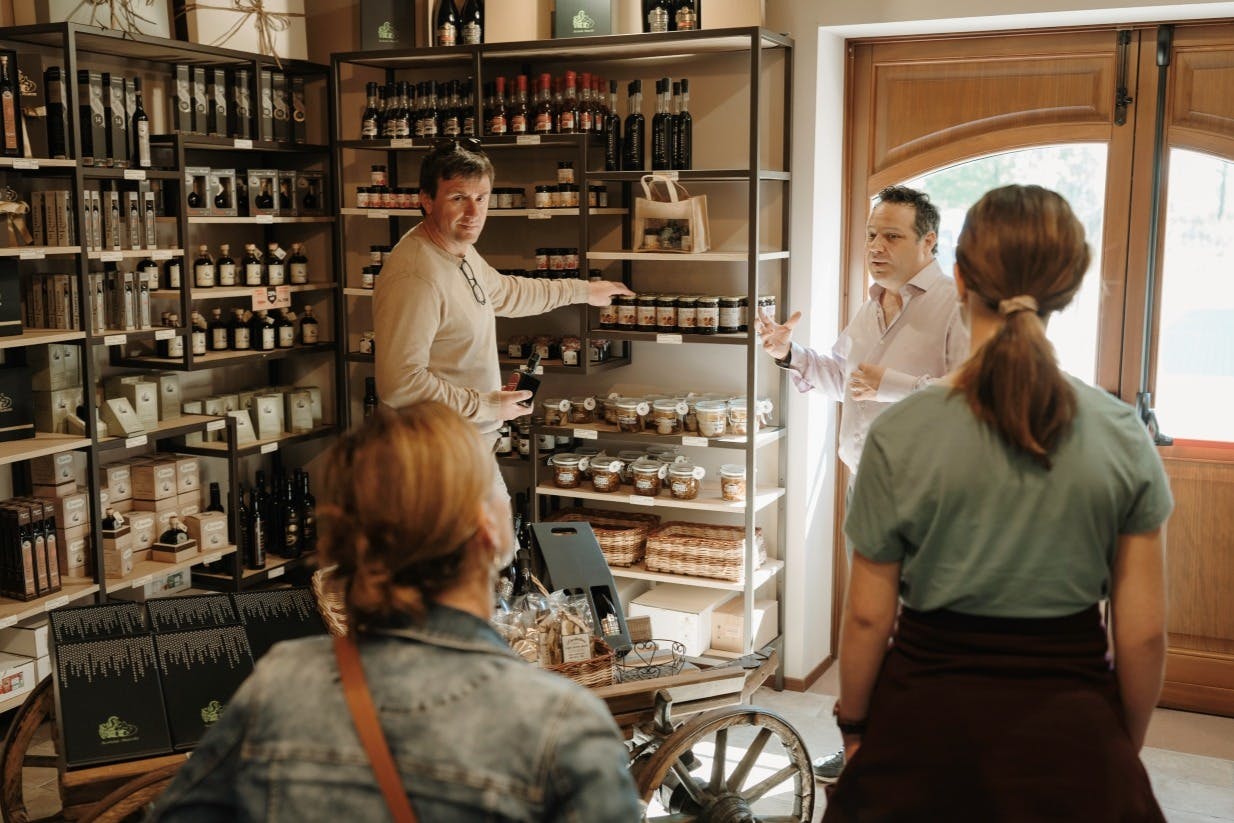 V obchodě stojí u polic se sklenicemi a lahvemi čtyři lidé. Jeden muž ukazuje na produkt, zatímco druhý vysvětluje.