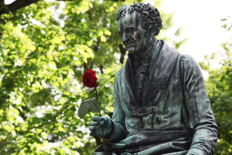 A weathered bronze statue of a person holding a red rose against a backdrop of green leafy trees.