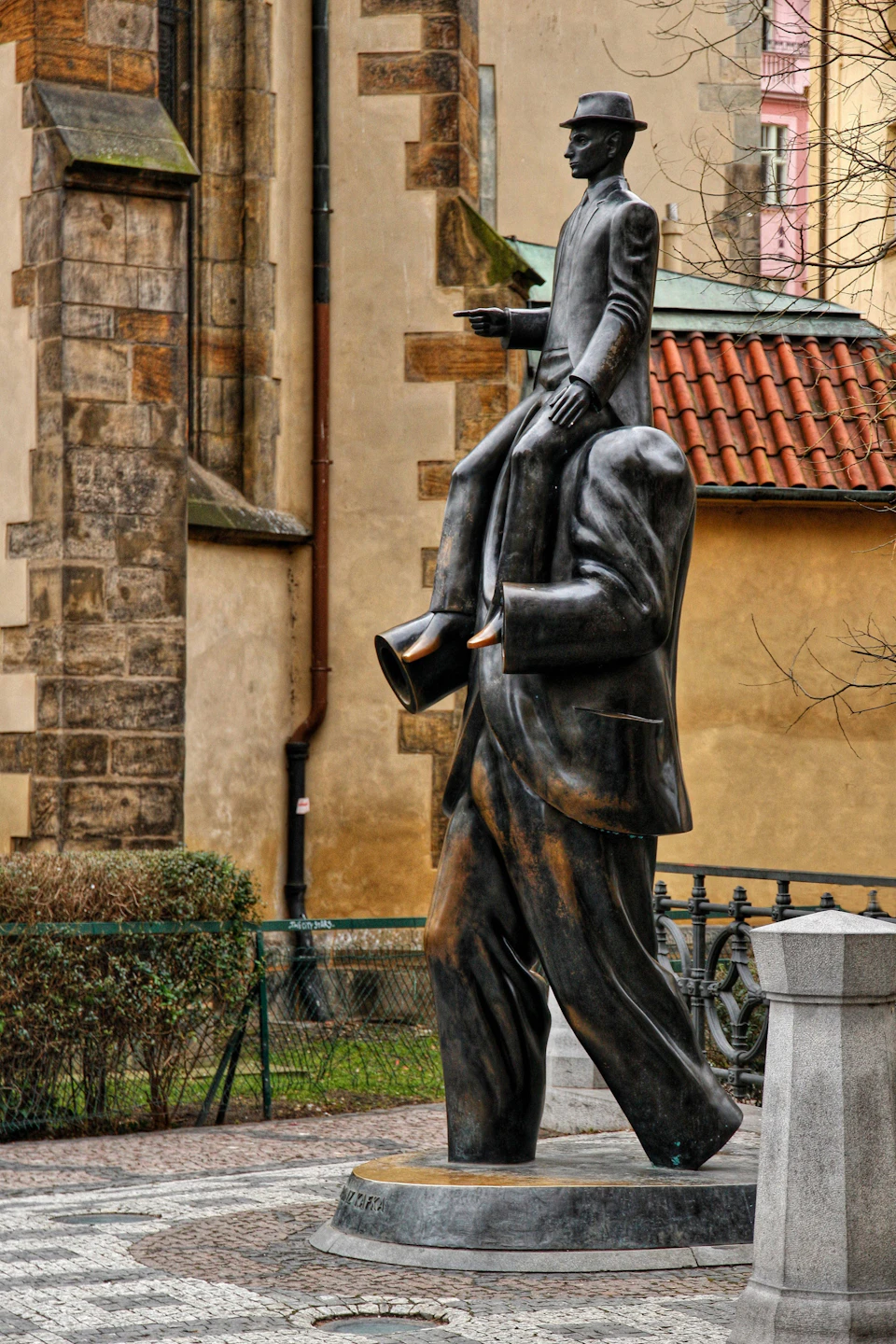 Bronze statue of a headless man carrying a smaller man on his shoulders, located outdoors near an old building.