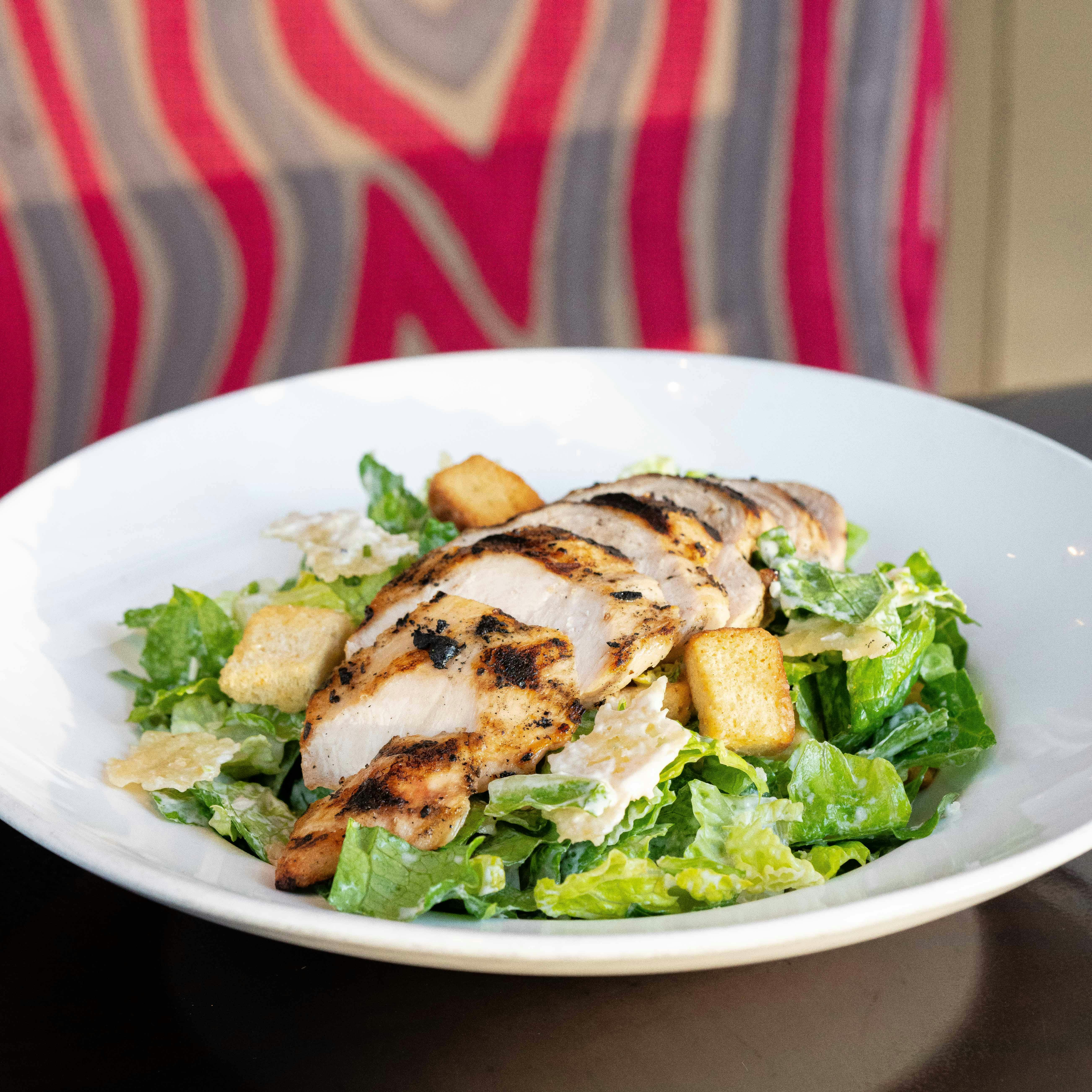 A Caesar salad topped with grilled chicken and croutons, served in a white bowl on a table with a patterned background.