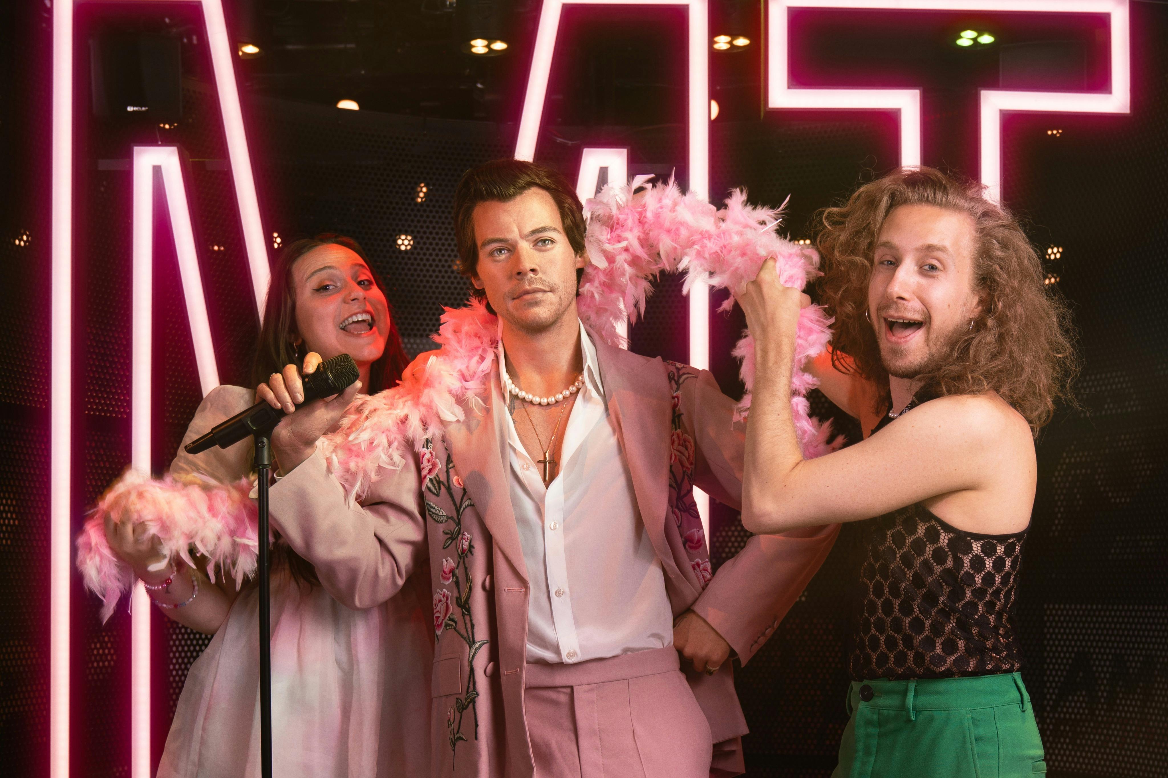 Τρεις άνθρωποι ποζάρουν μαζί, με την κεντρική φιγούρα να φορά ένα ροζ κοστούμι και έναν φτερωτό βόα, πλαισιωμένο από λαμπερά φώτα νέον.