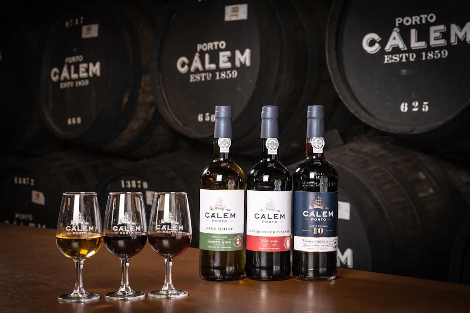 Three bottles of Calem port wine and four glasses on a table, with barrels labeled "Calem" in the background.