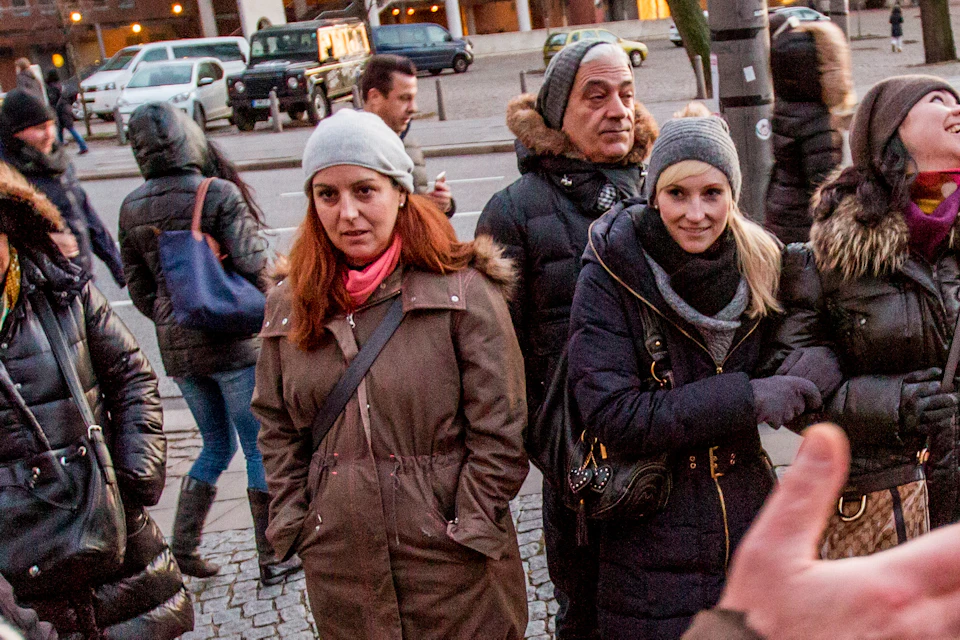 A group of people dressed in winter clothing gather outdoors near buildings, listening to a person speaking animatedly.