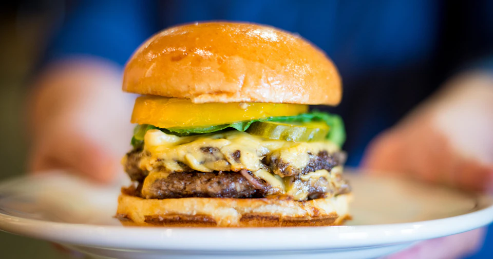 A cheeseburger with a shiny bun, melted cheese, lettuce, pickles, and tomato slices on a white plate.