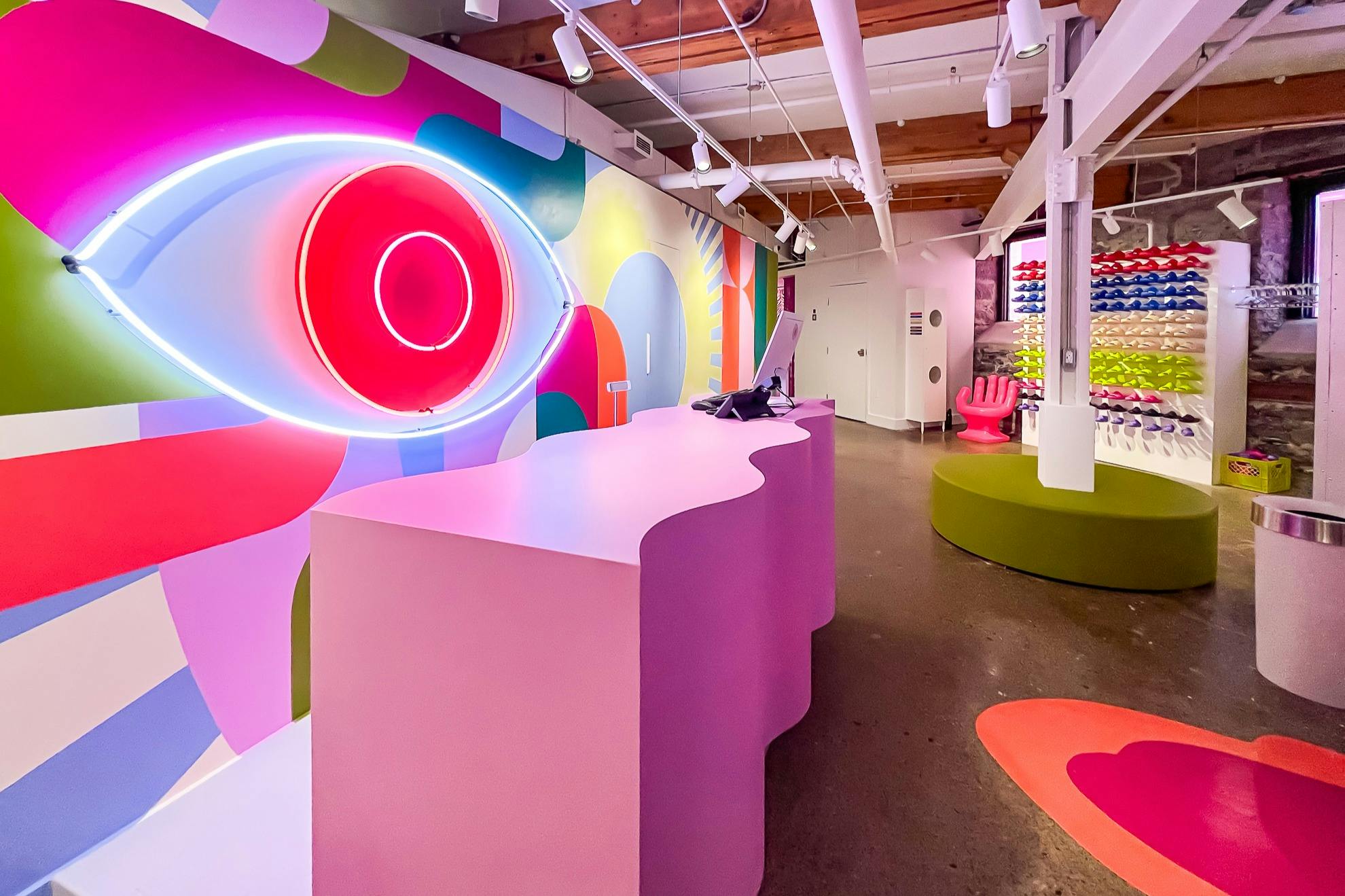 A colorful interior with a wavy pink desk, a neon eye-shaped light, and a display of colorful sunglasses on the wall.