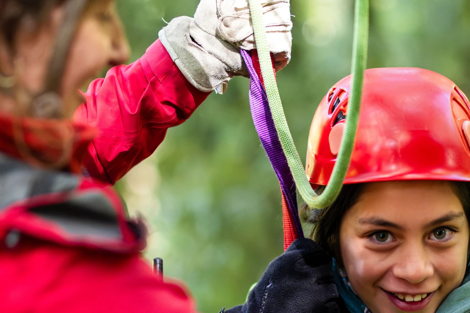 Two individuals in helmets and harnesses engaging in an outdoor activity in a forested area, one assisting the other.