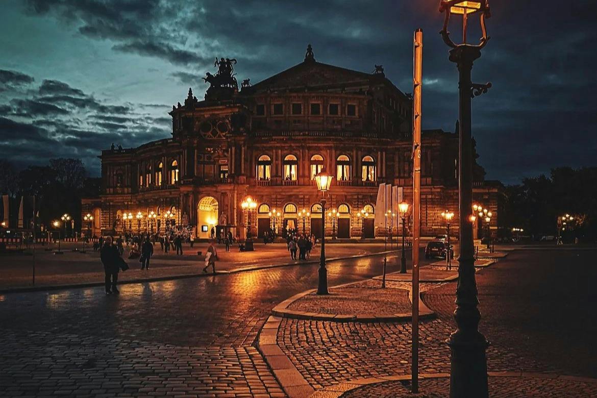 Un edificio storico illuminato di notte dai lampioni, con persone che passeggiano su una piazza acciottolata sotto un cielo nuvoloso.