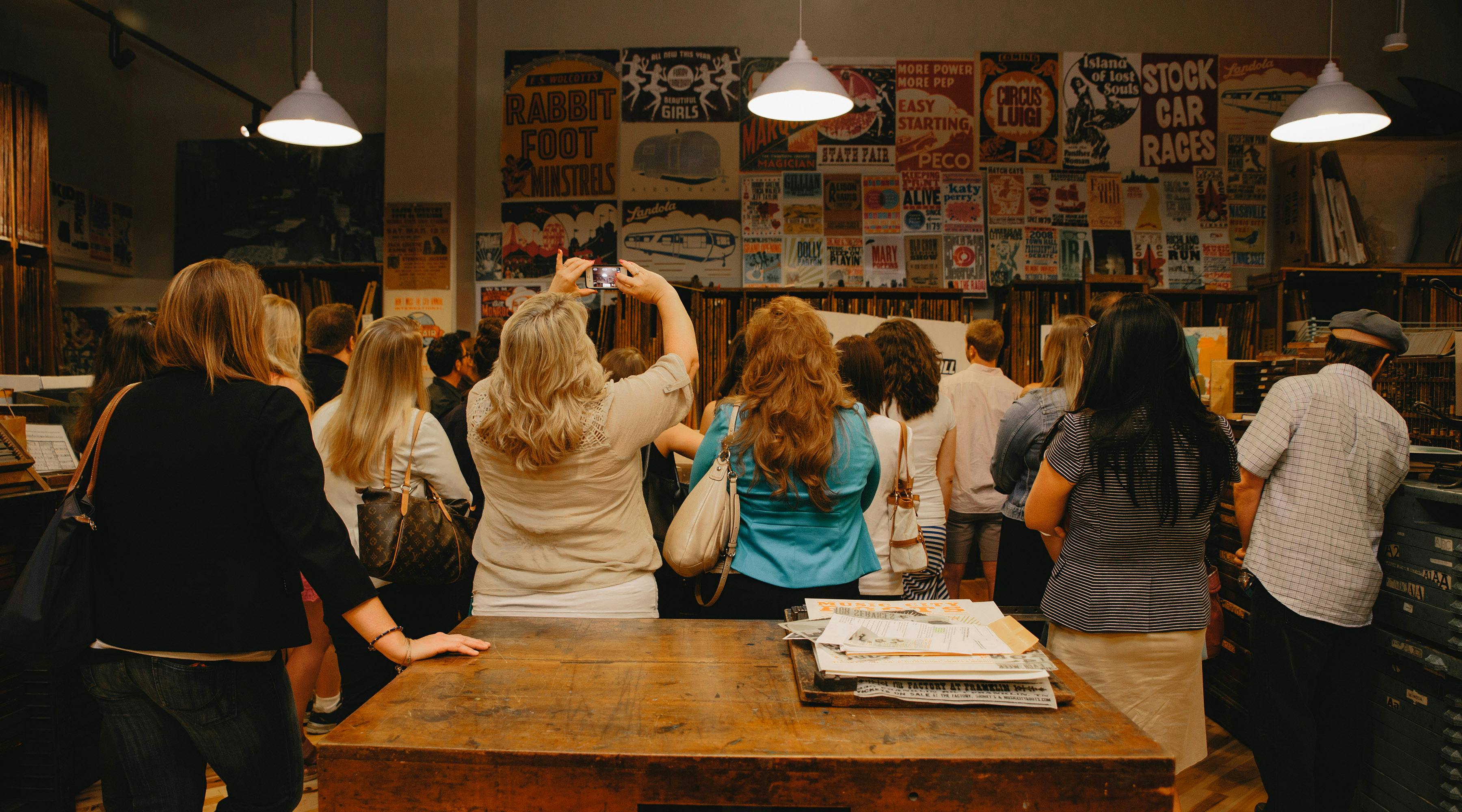 A group of people gathered in a room with vintage posters on the wall; one woman is taking a photo with her phone.
