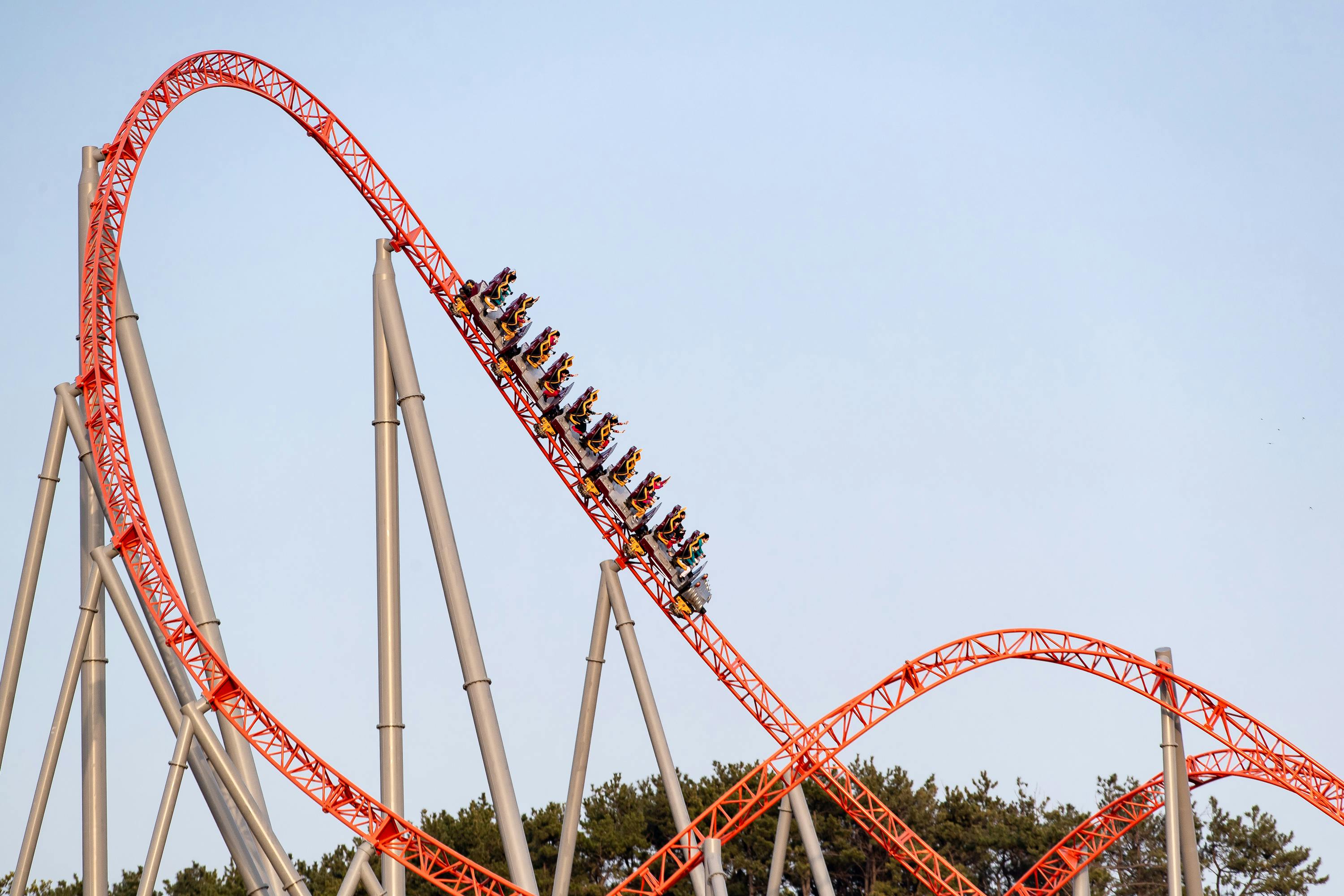 A roller coaster with a red track and a car full of riders descending a steep drop against a clear blue sky.