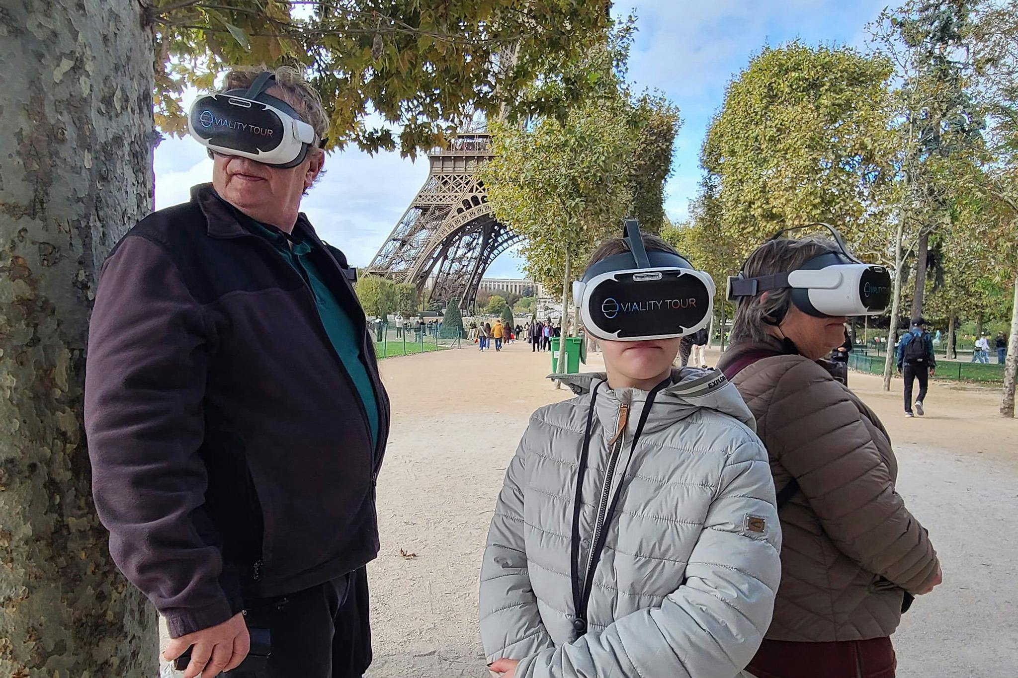 Tre persone che indossano cuffie VR si trovano vicino alla Torre Eiffel, circondate da alberi e da un cielo limpido.