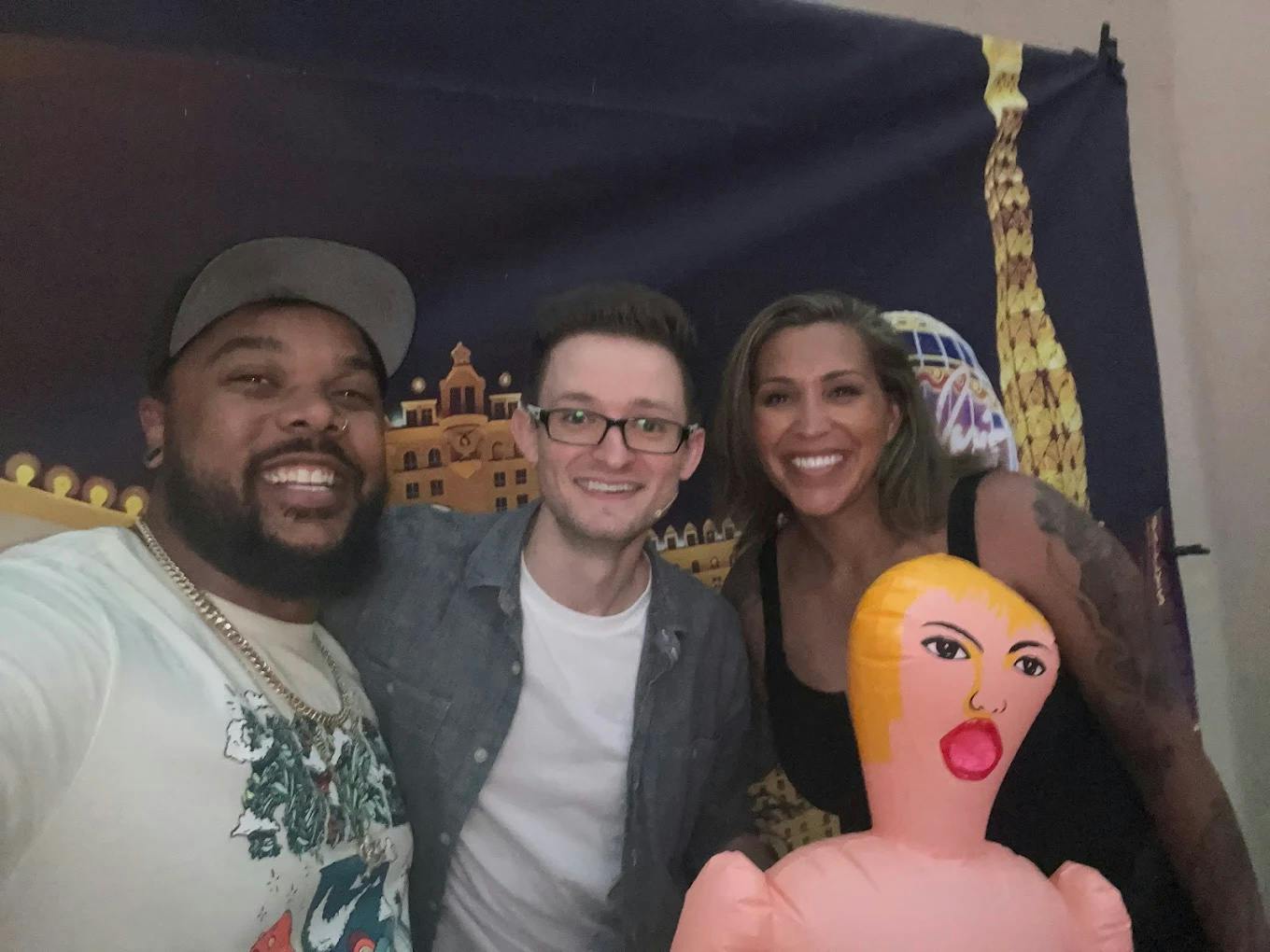 三个人对着镜头微笑，前面是一个充气娃娃，背景描绘了城市景观。