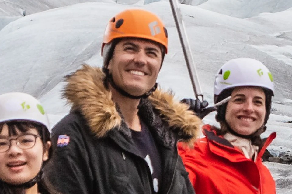 A group of people wearing helmets and outdoor gear stand on a glacier with ice axes, with an icy landscape in the background.