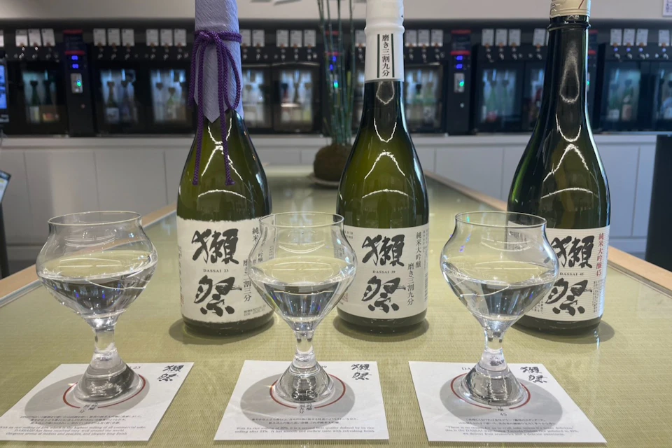 Three glasses of clear liquid in front of three sake bottles on coasters, set on a table with a modern background.