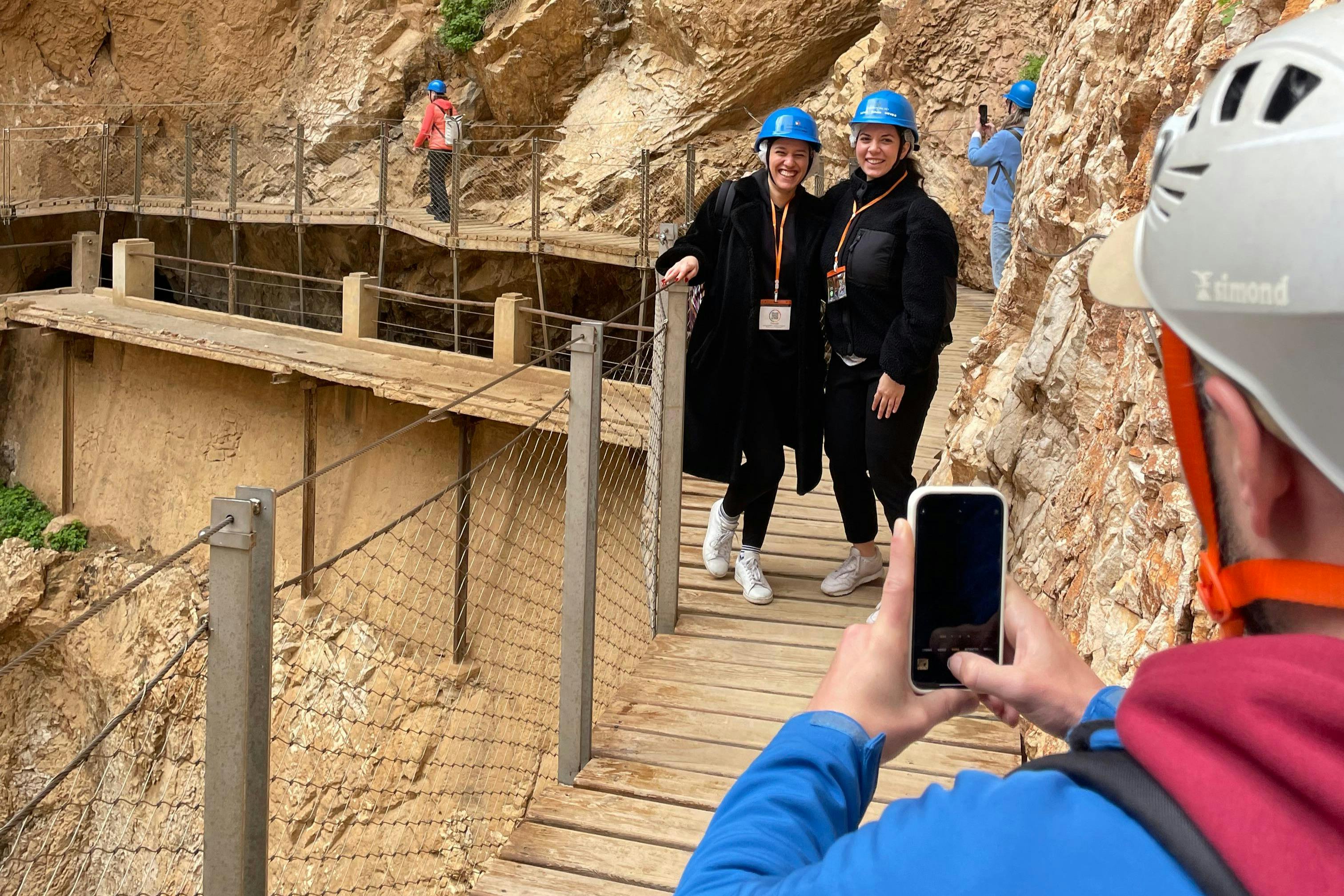 Two people in blue helmets pose for a photo on a rocky, wooden walkway while another person takes their picture.