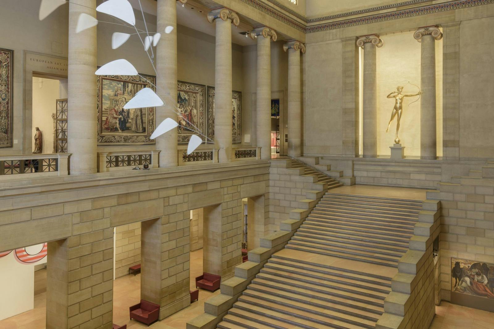 Una gran escalera en un museo con columnas, tapices, una escultura móvil y una estatua al fondo.