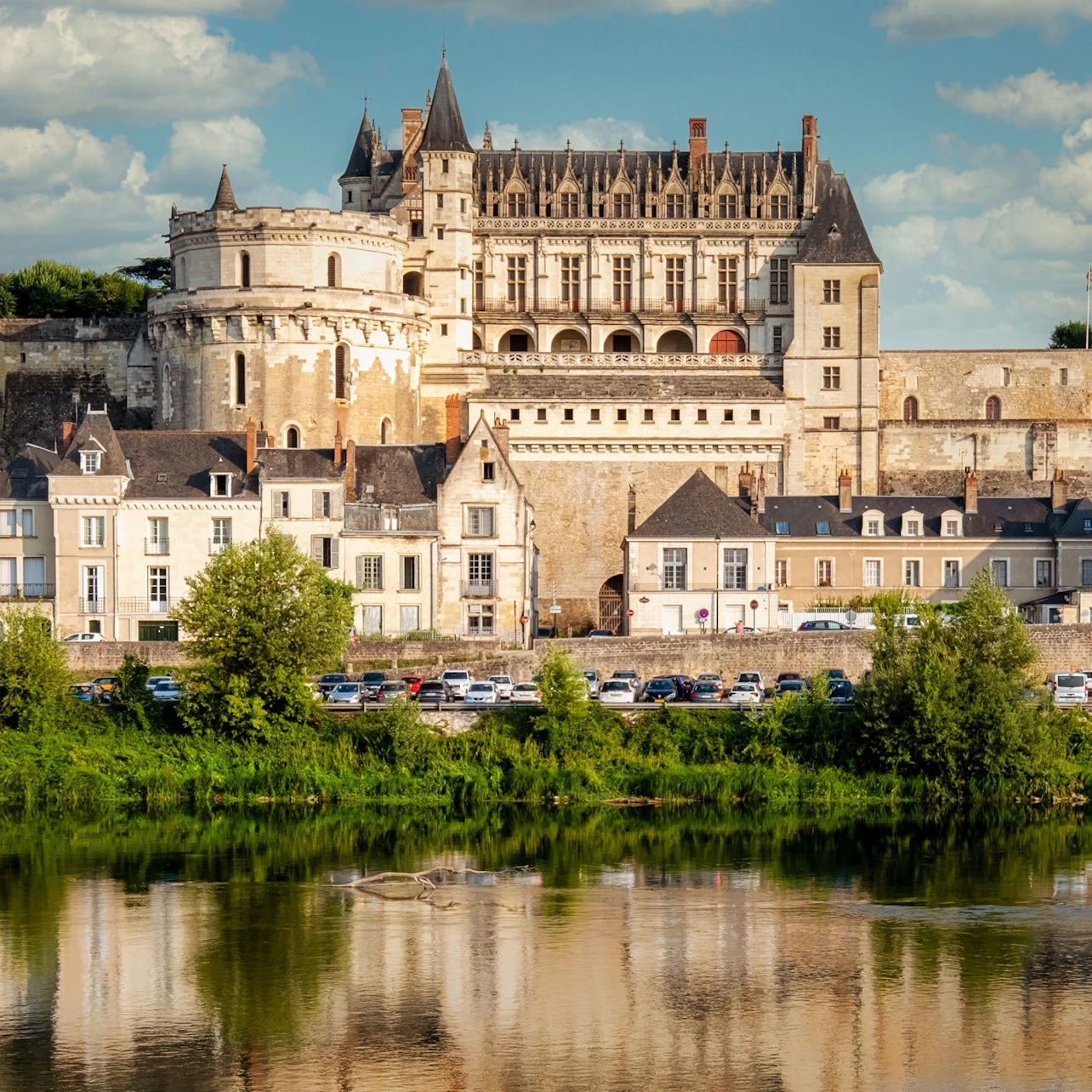 Un château historique au sommet d'une rangée de bâtiments, se reflétant dans une rivière, avec de la verdure et un ciel bleu en arrière-plan.