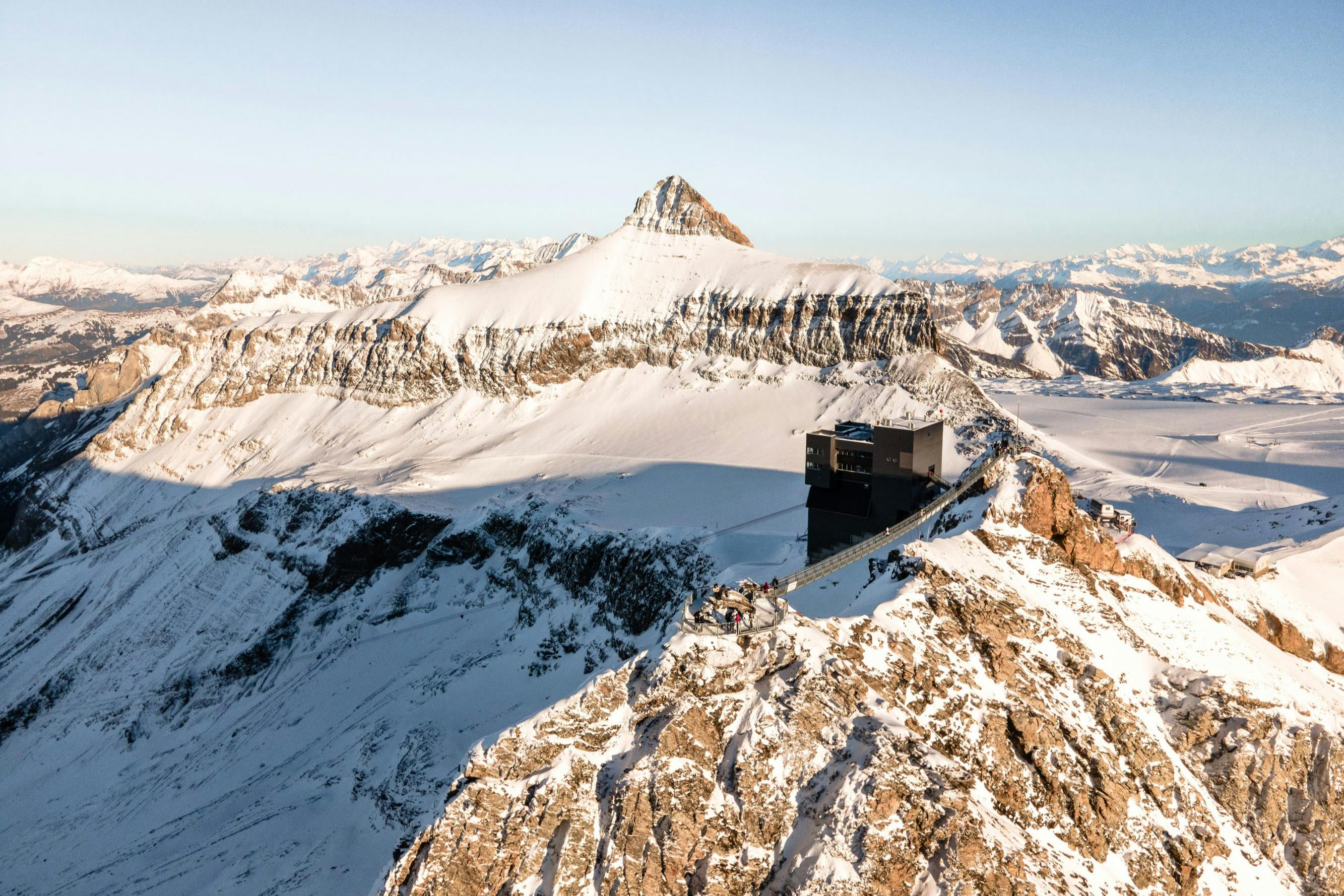 봉우리를 배경으로 눈 덮인 산 풍경과 전경의 바위 능선에 자리한 현대적인 건물.