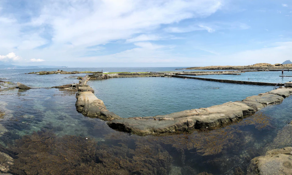 A natural tidal pool surrounded by rocky edges with seaweed visible underwater, set against an ocean and sky background.