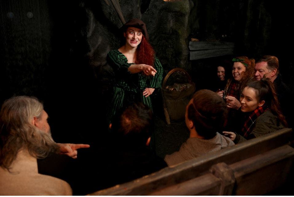 A woman in a green dress and hat is entertaining a seated group of people in a dimly lit, rocky setting.