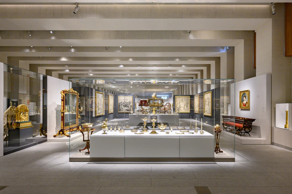 A museum exhibit displaying ornate tableware and artifacts in a glass case, surrounded by framed artworks on the walls.