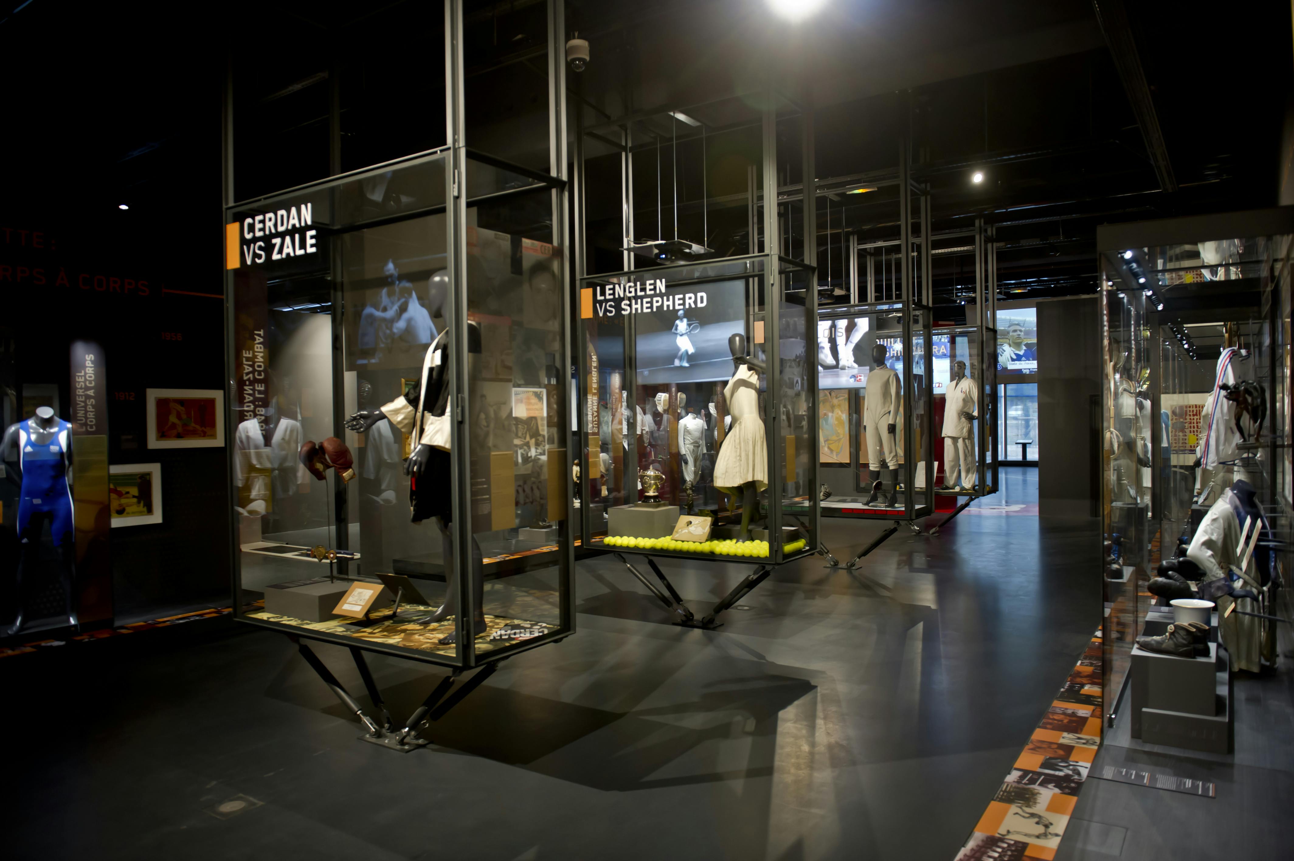 Expozice muzea s figurínami s historickým sportovním oblečením ve skleněných vitrínách označených „CERDAN VS ZALE“ a „LENGLEN VS SHEPHERD“.