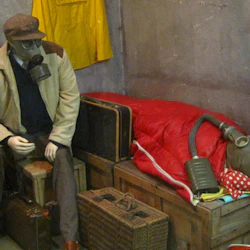 Skyltdockor med gasmasker sitter bland resväskor och en säng i ett rum med grå väggar, gul kappa och inramade bilder.
