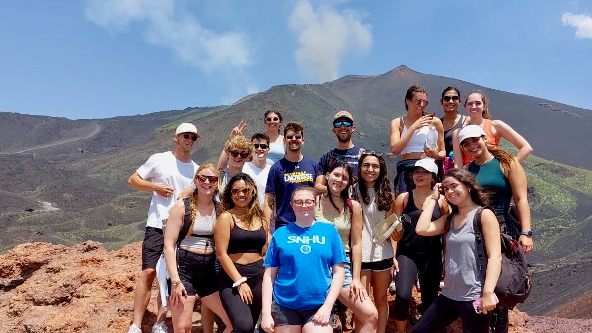 Μια ομάδα 16 ατόμων ποζάρει μπροστά από ένα ηφαίστειο που καπνίζει κάτω από έναν καταγάλανο ουρανό.