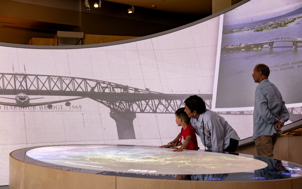 Three people interact with an exhibit featuring a curved screen displaying historical bridge images and a map.