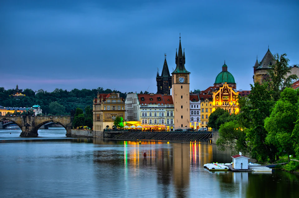 Historic buildings with towers and domes along a riverfront, under a cloudy evening sky, with reflections in the water.